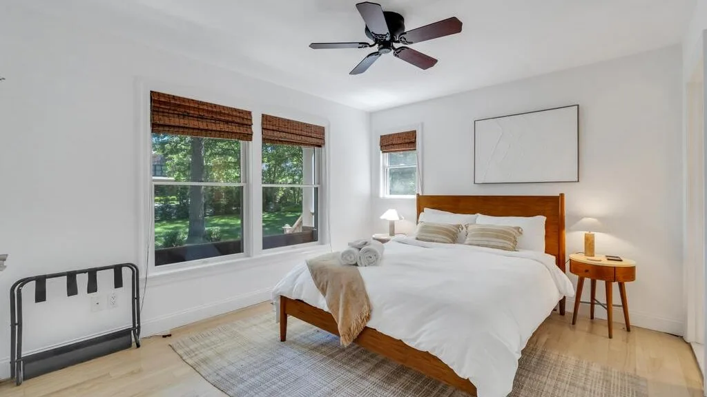 Un dormitorio luminoso con cama de madera, ropa de cama blanca y ventanas con vistas a la vegetación.