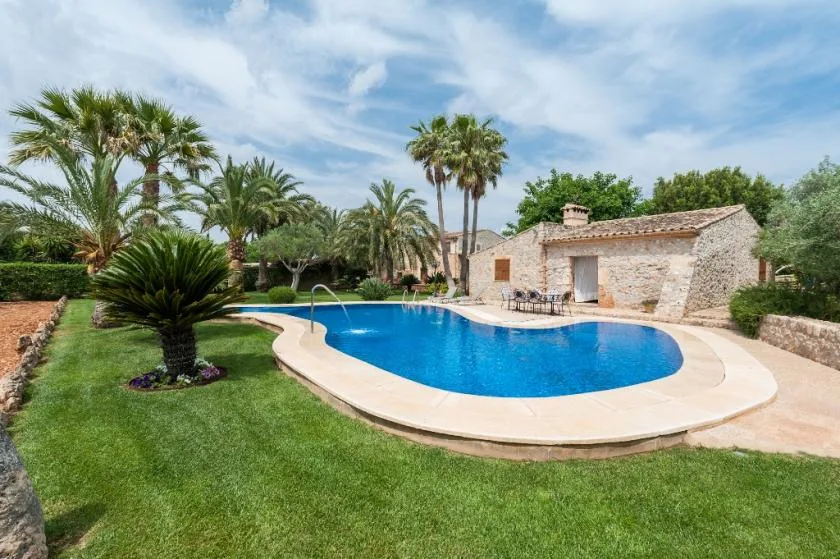 A curved swimming pool sits in a landscaped garden beside a stone building.