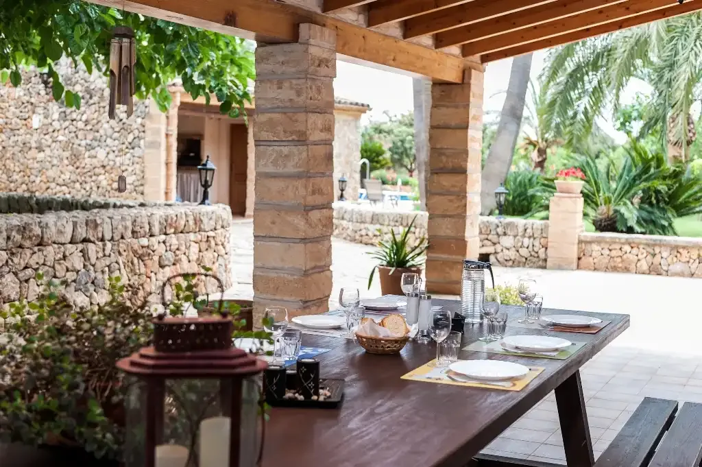 Un comedor en un patio cubierto, con una mesa junto a muros de piedra y vegetación.