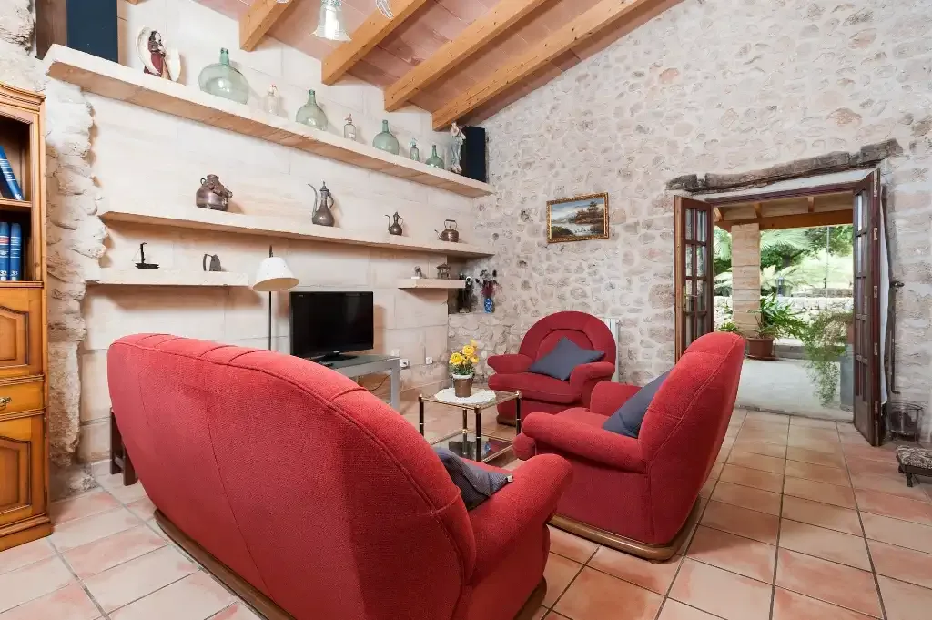 Una sala de estar con paredes de piedra tiene asientos rojos, un televisor y puertas abiertas a un patio.