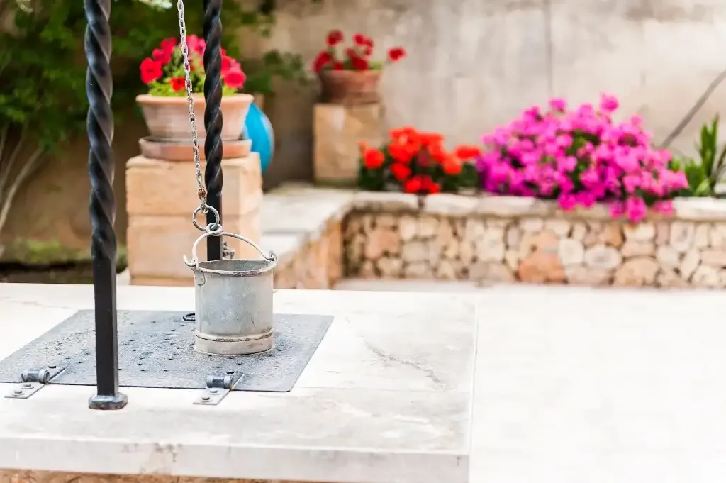 Un pozo de piedra con un cubo colgante está junto a un jardín lleno de flores.