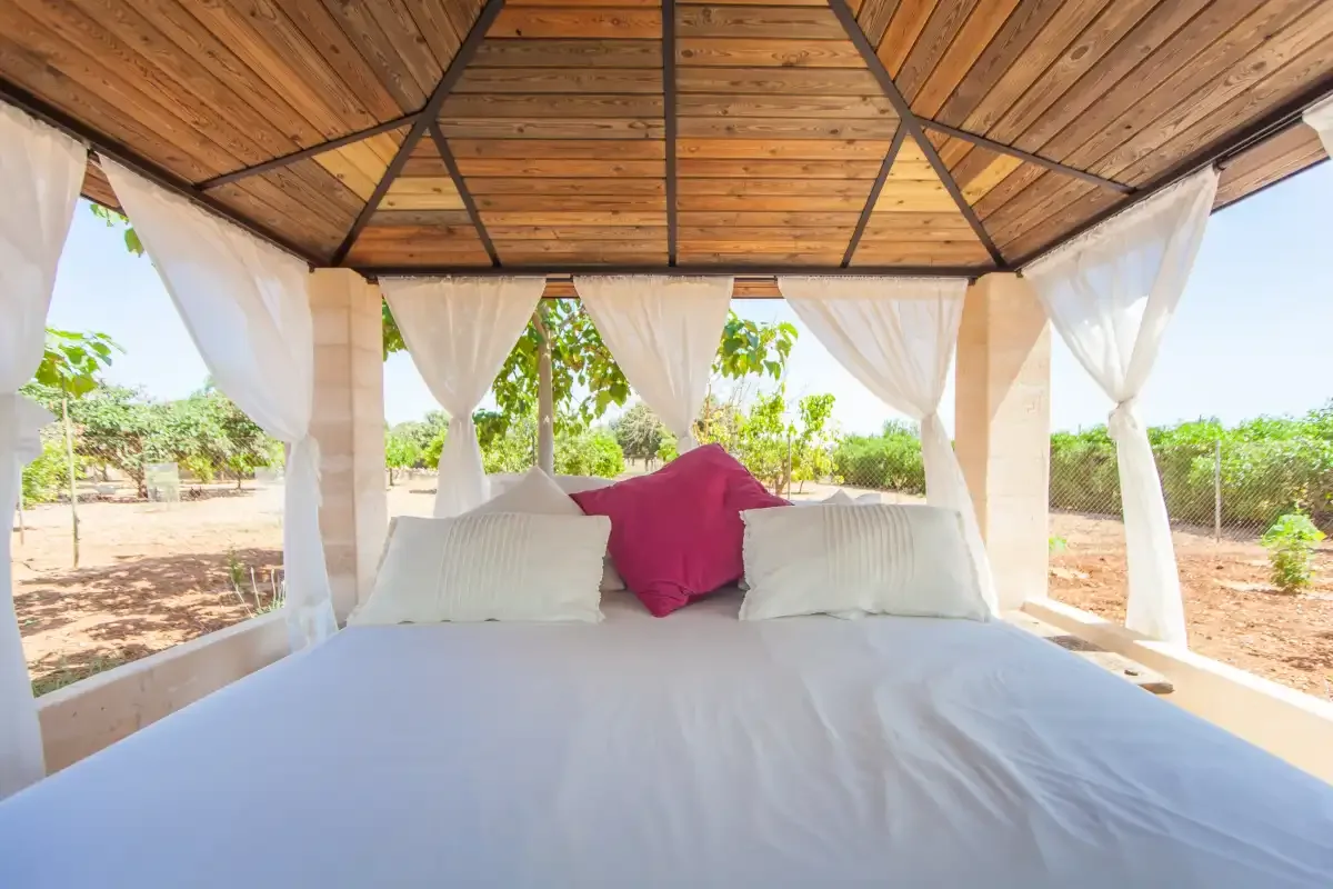 Una cama al aire libre está bajo un techo de madera con cortinas transparentes.