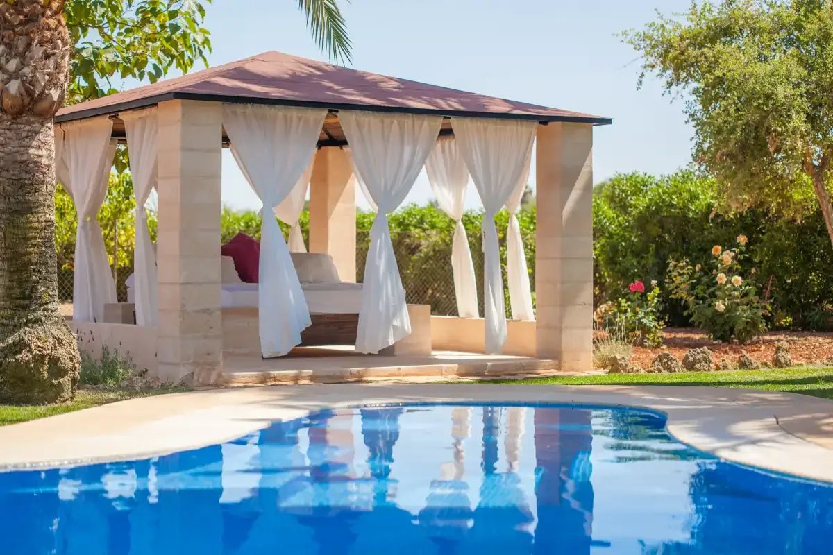 Una piscina de jardín está junto a un pabellón con cortinas y asientos.