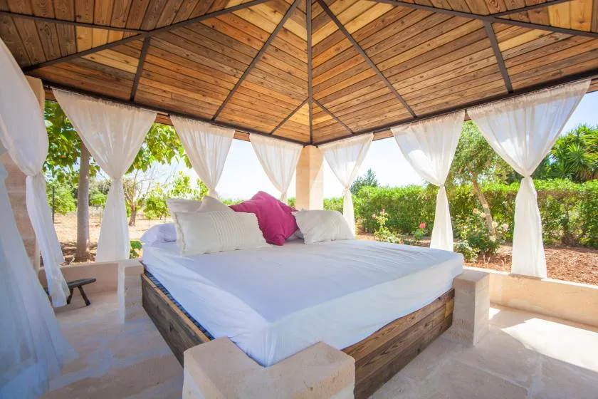 A large bed sits beneath a wooden pavilion roof with white curtains.