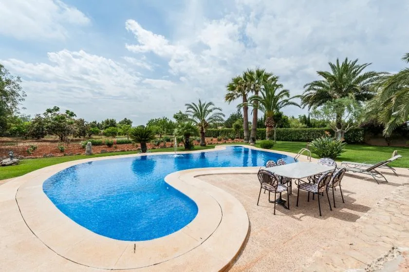 A curved swimming pool sits beside an outdoor dining table in a landscaped garden.