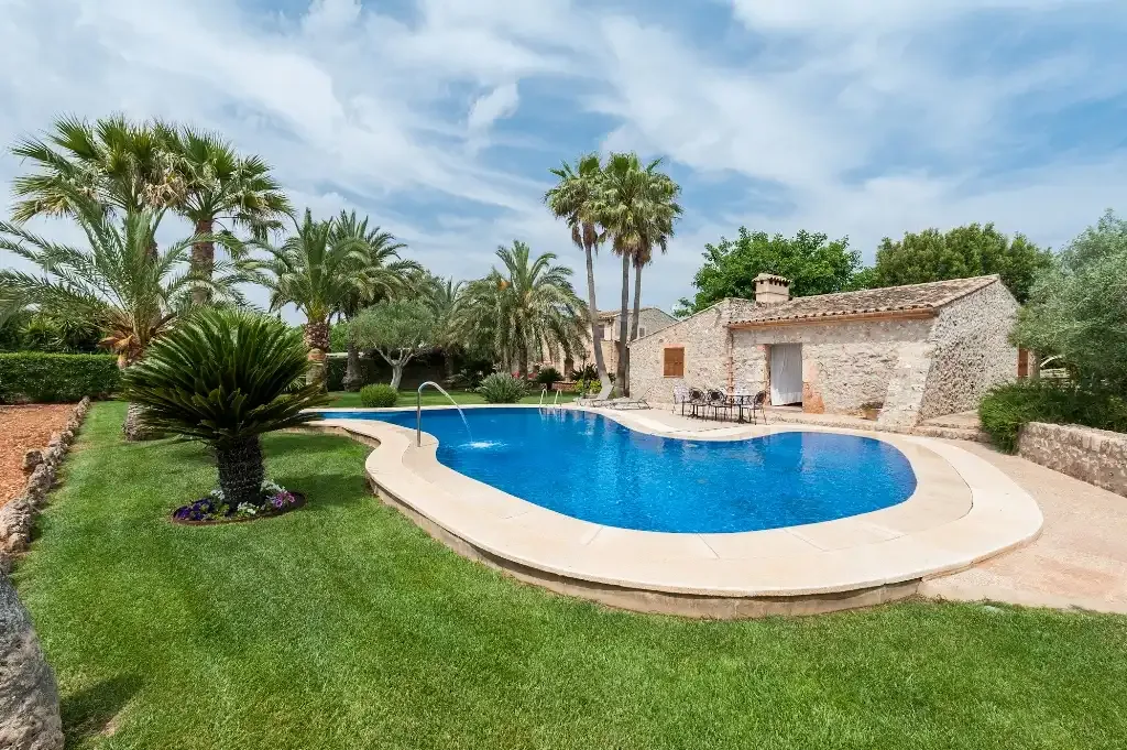 Una piscina curva atraviesa un césped con palmeras junto a un edificio de piedra.