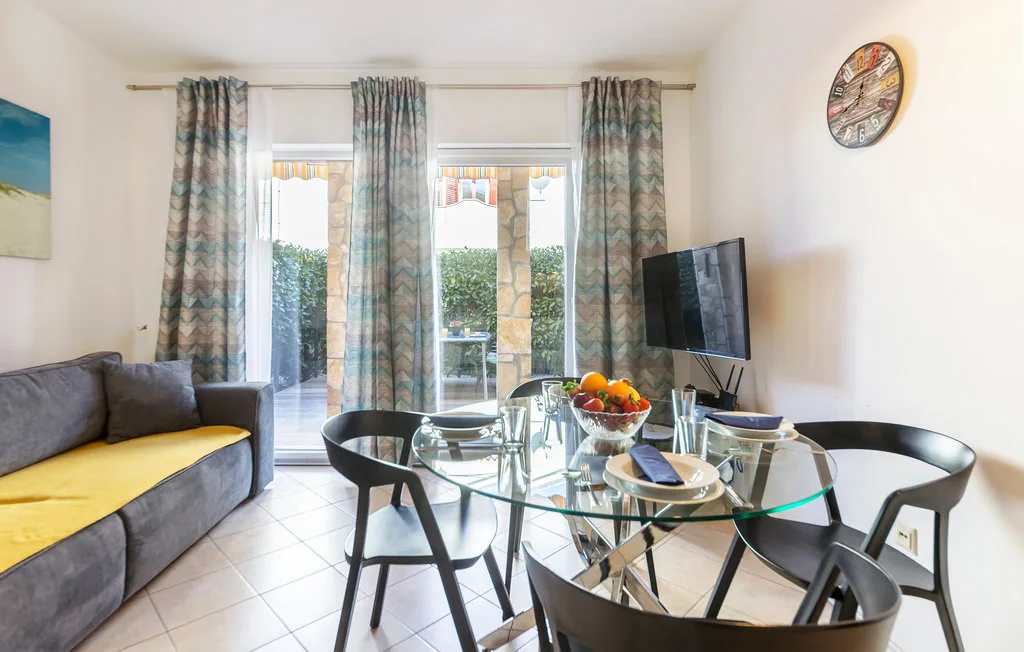 Una luminosa sala de estar y comedor tiene una mesa de vidrio y asientos junto a ventanas con cortinas.