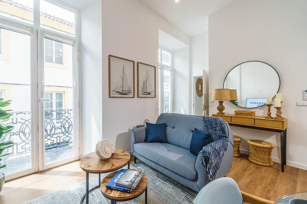 Una sala luminosa con un sofá azul junto a ventanas altas y una consola de madera.