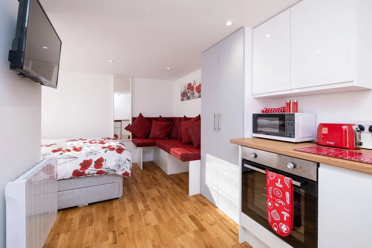 A compact studio room with a bed, red seating, and a kitchenette.