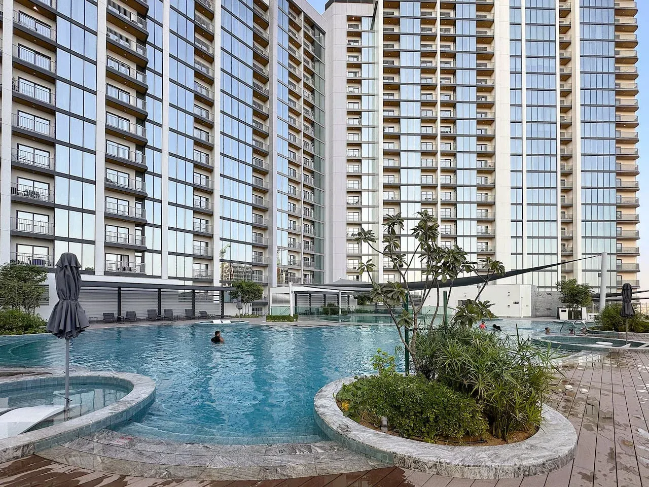 A swimming pool sits among tall residential towers, with landscaped planting along the water.