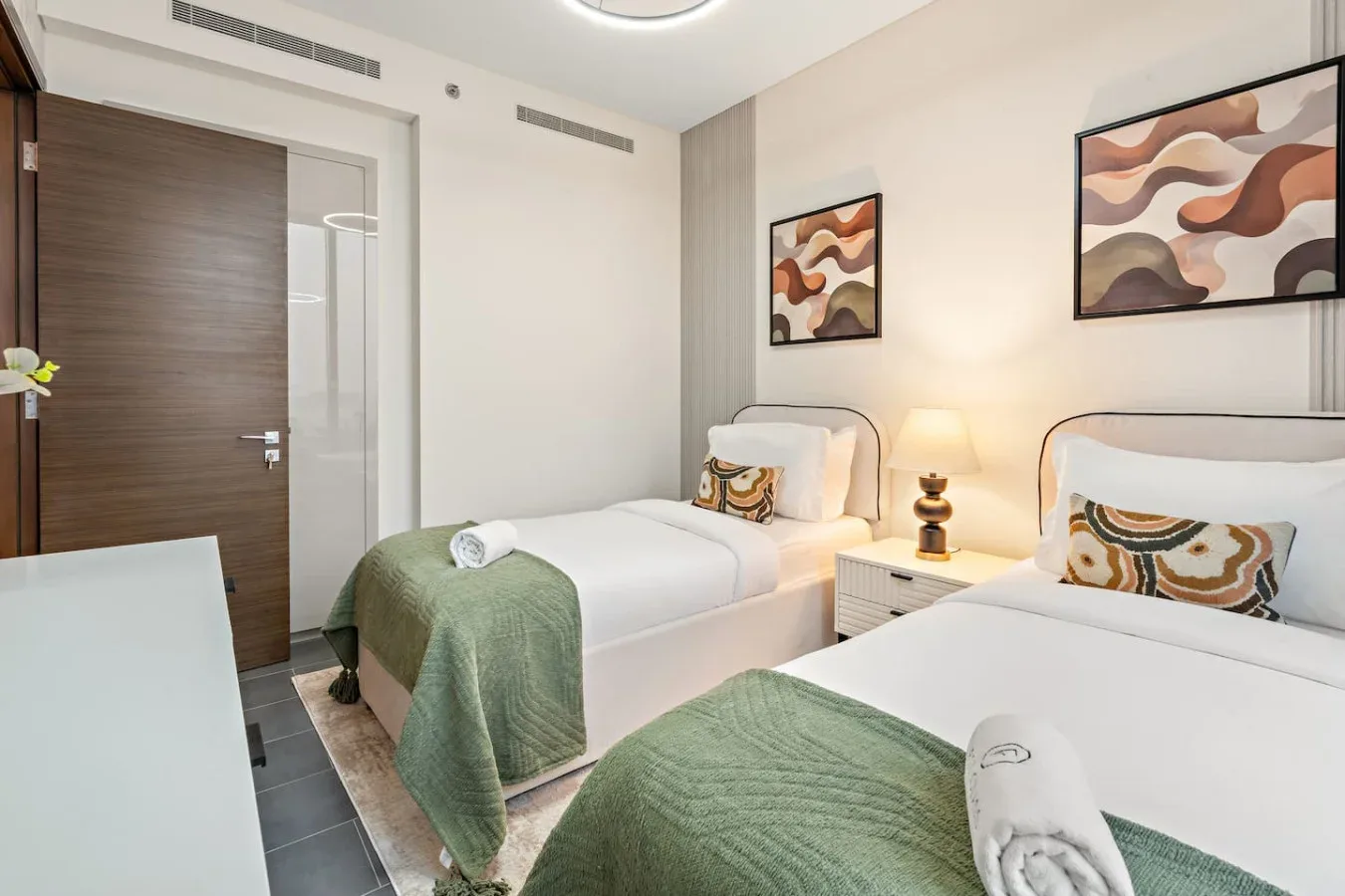 A bright bedroom with two beds, green blankets, and abstract wall art.