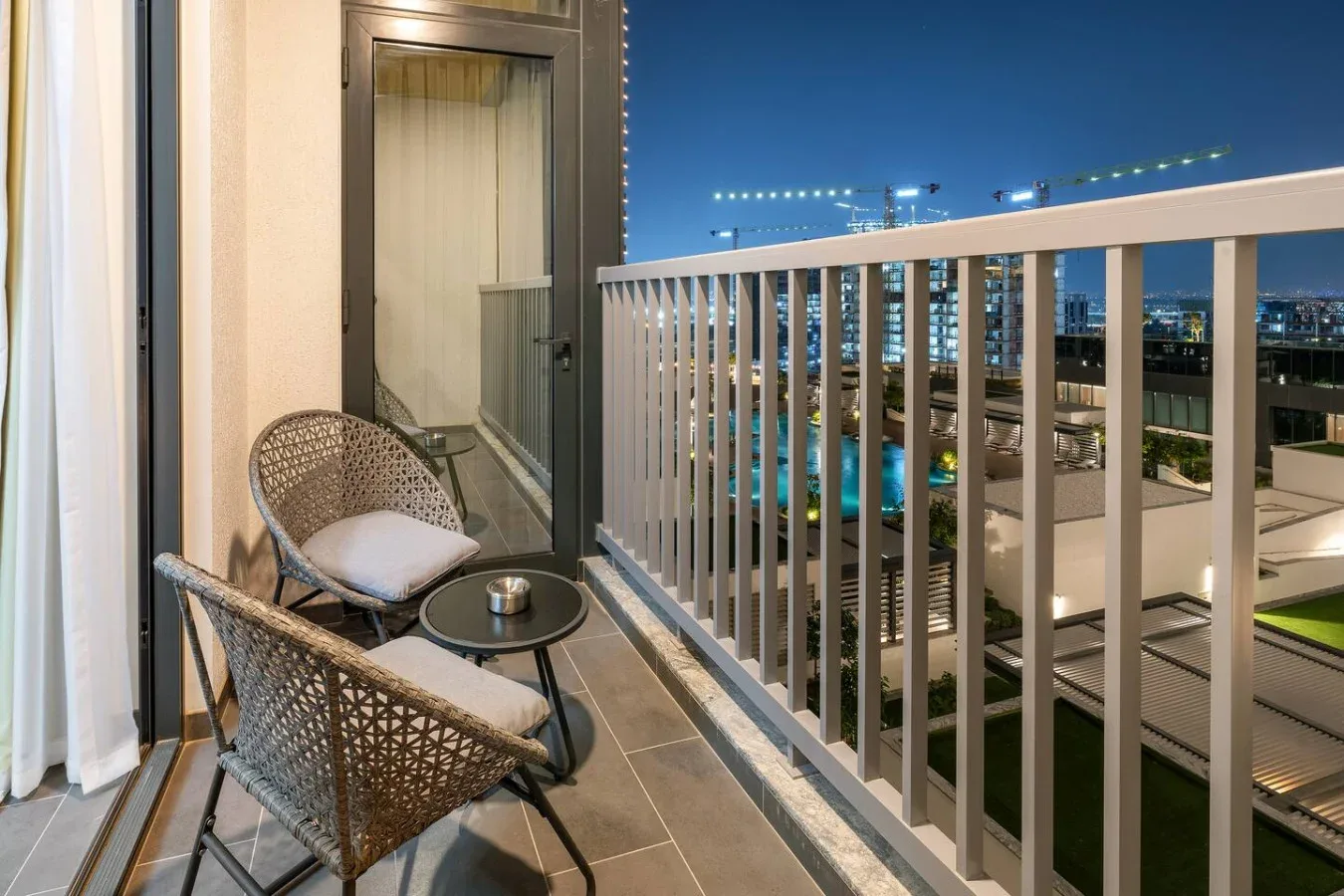 A balcony with wicker seating overlooks a brightly lit city at night.