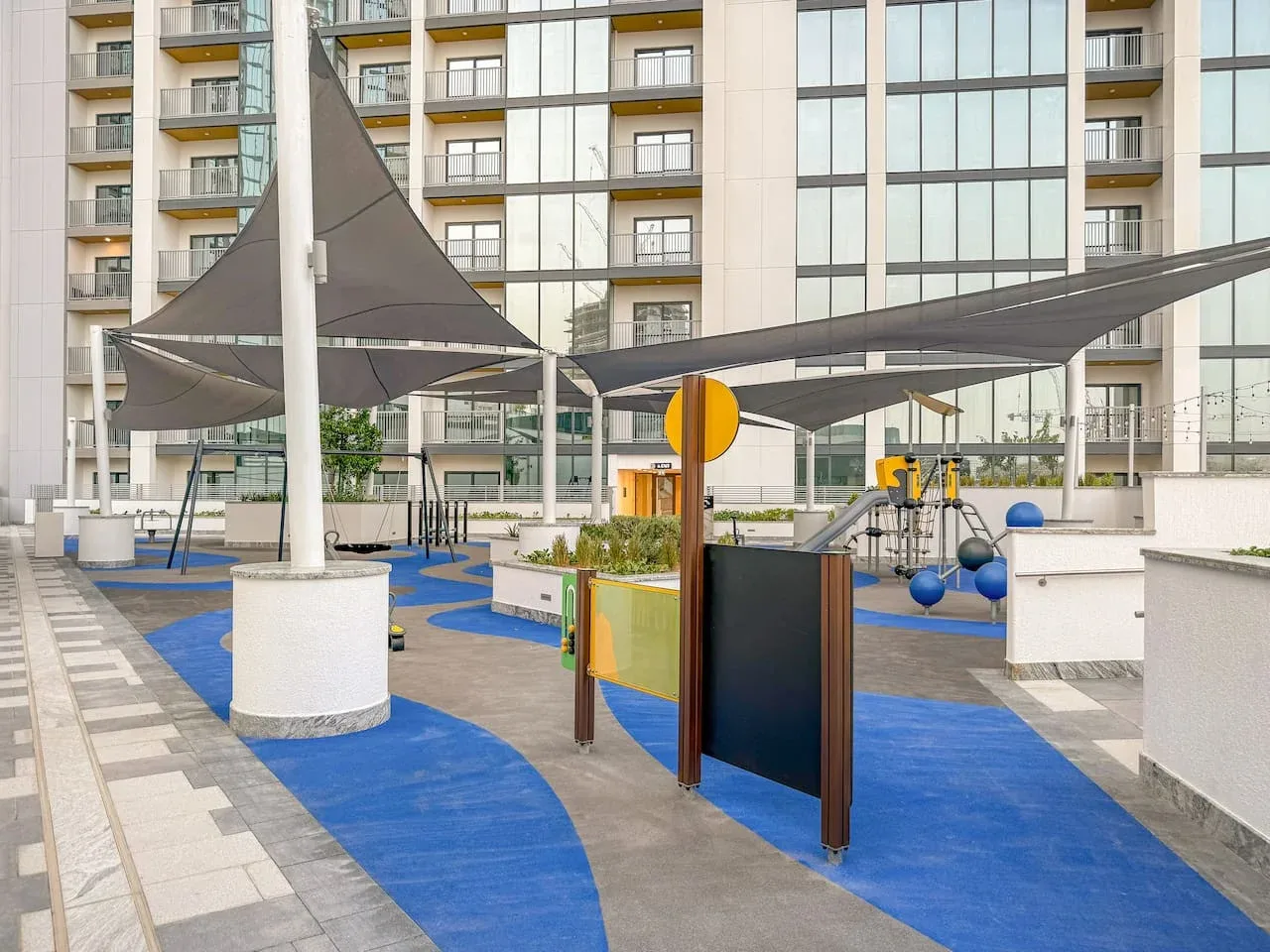 A shaded outdoor play area with colorful playground equipment beside apartment buildings.