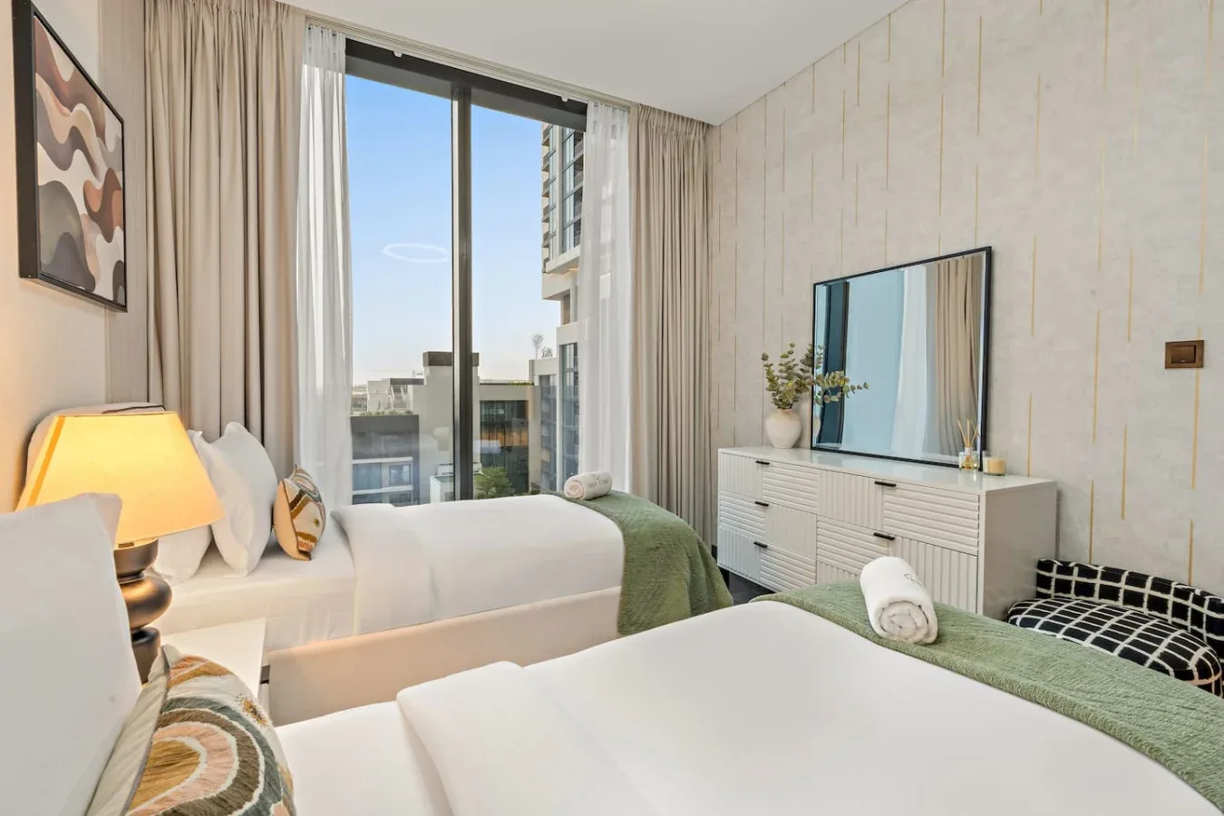 A bright bedroom with two beds, floor-to-ceiling curtains, and a dresser.