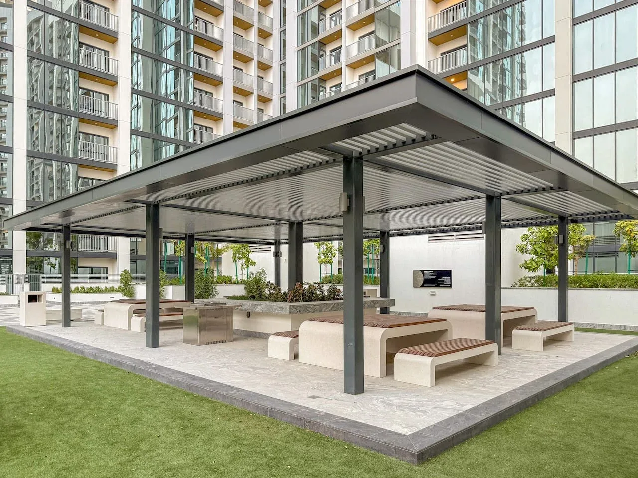A covered outdoor seating area with benches and tables beside a lawn.