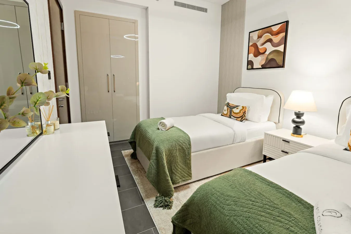 A bright bedroom with two beds, green blankets, and bedside lighting.