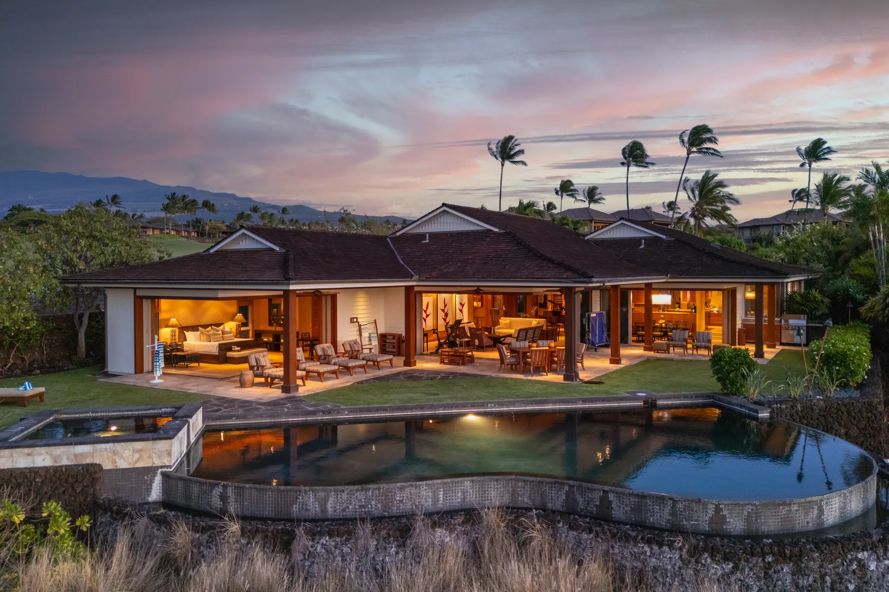 A modern home opens onto a curved swimming pool beneath a colorful evening sky.