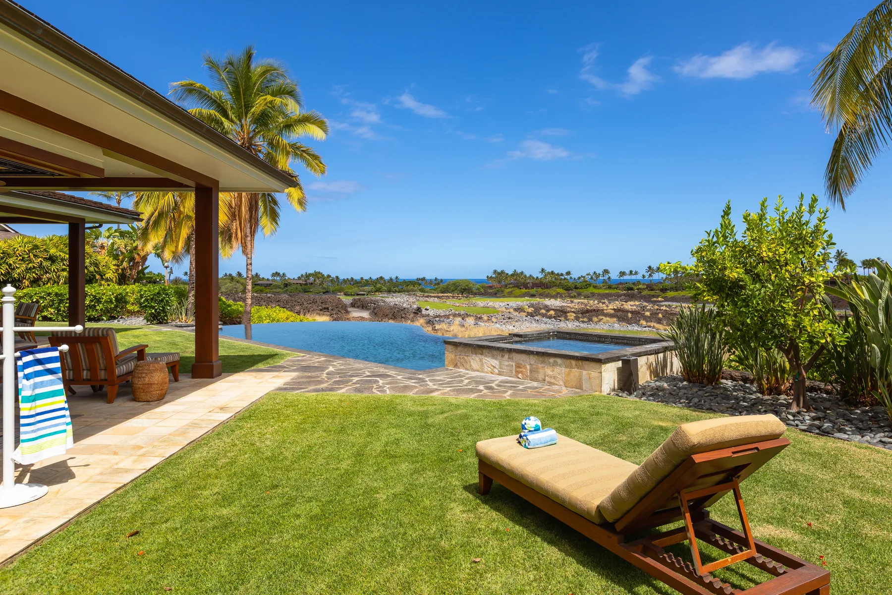 A sunny outdoor pool area with a lawn and reclining seating beside a covered patio.