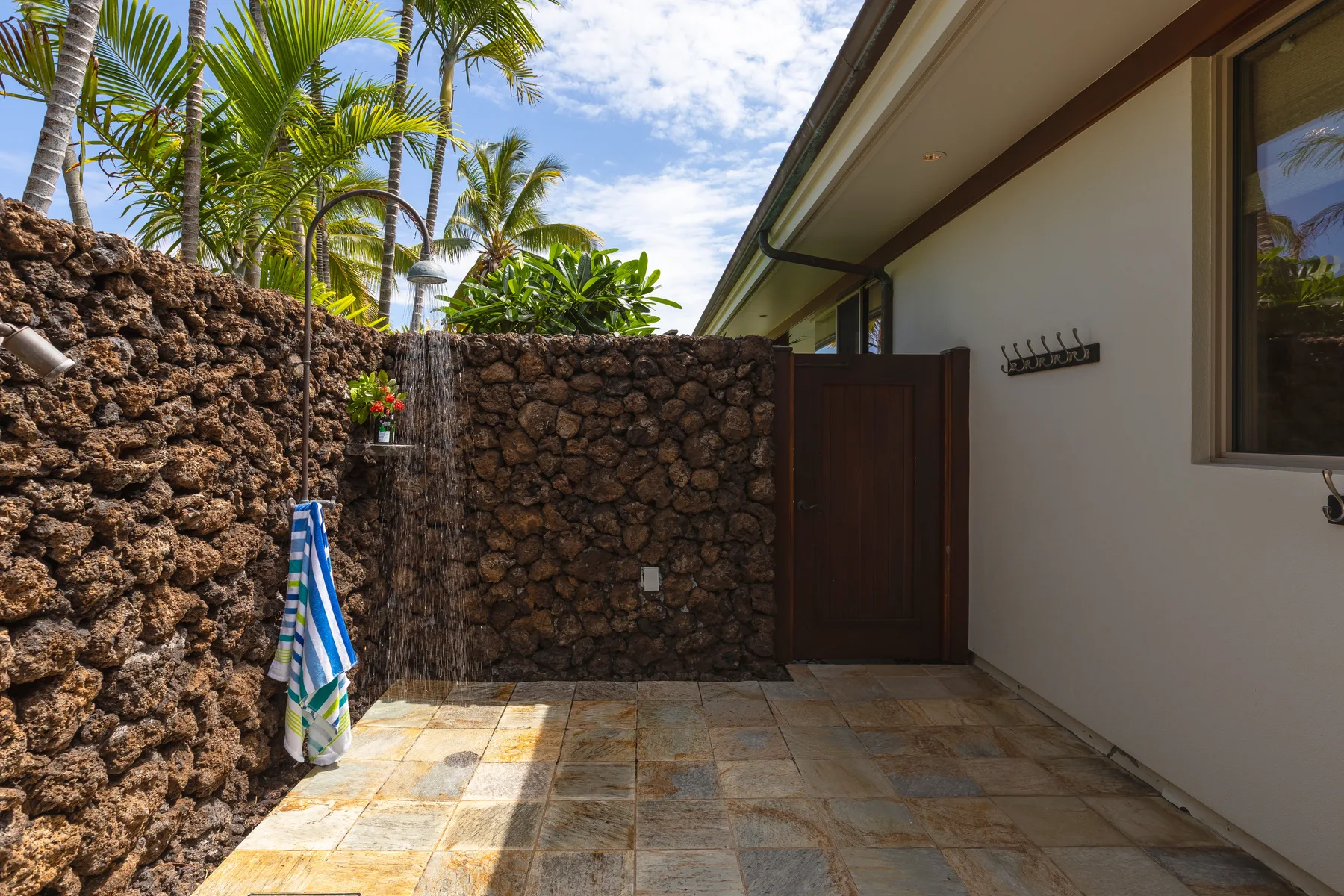 An outdoor shower area beside a stone wall, with tropical plants overhead.