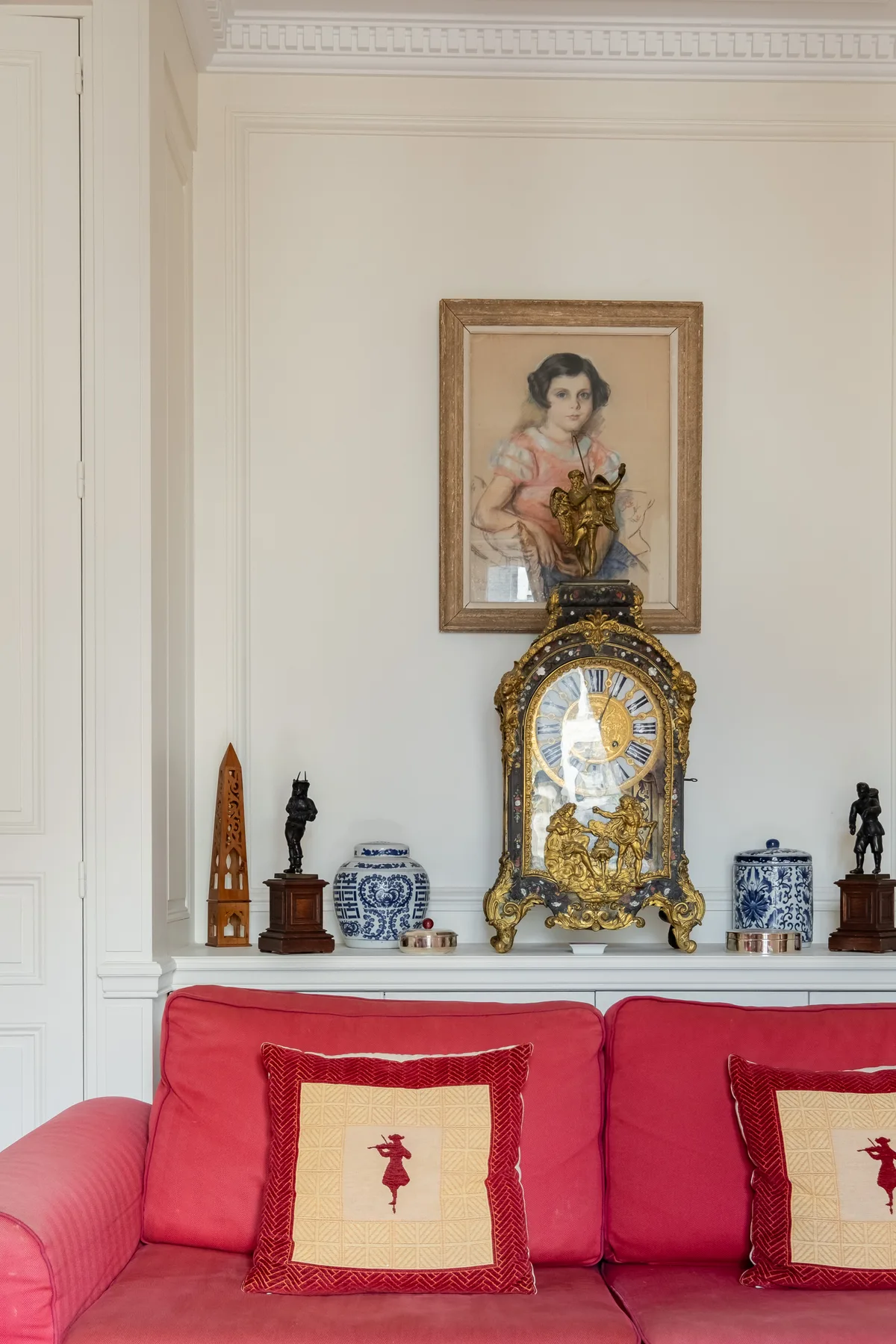 Un sofá rojo está bajo un reloj ornamentado y un retrato enmarcado en una sala luminosa.