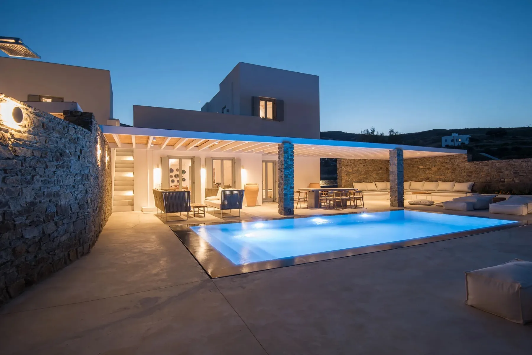 A modern house opens onto a lit swimming pool and covered outdoor seating area.