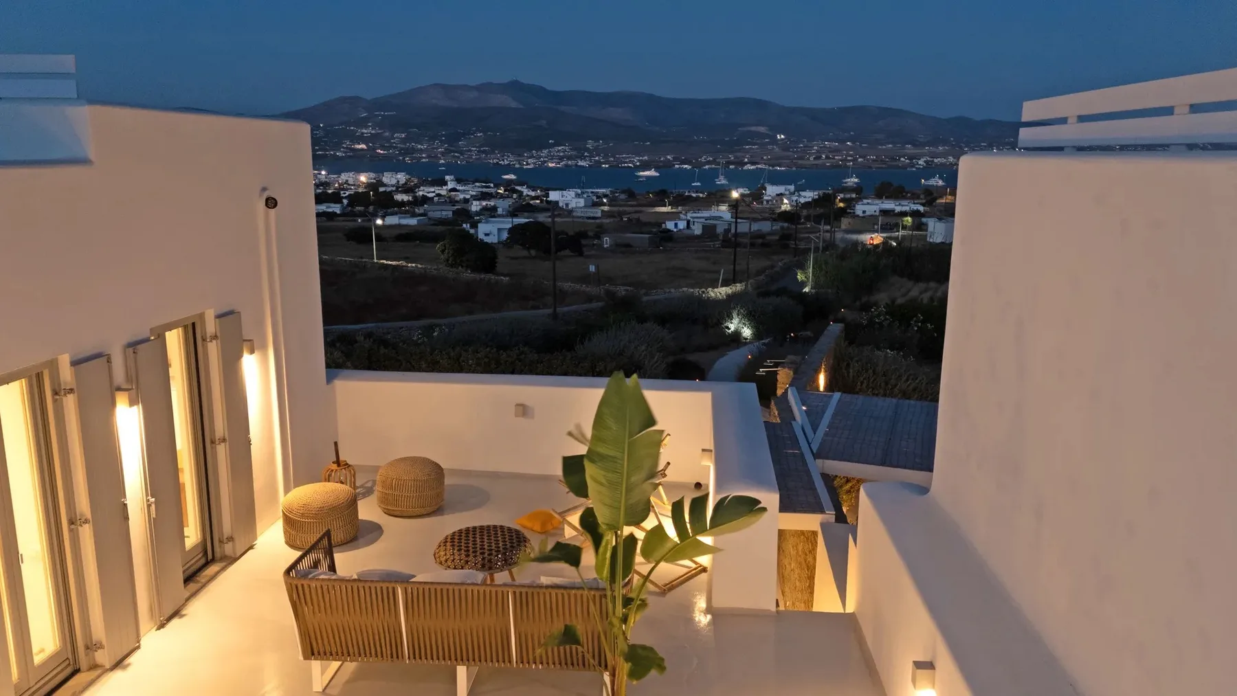 A rooftop terrace overlooks a coastal town and distant hills at dusk.