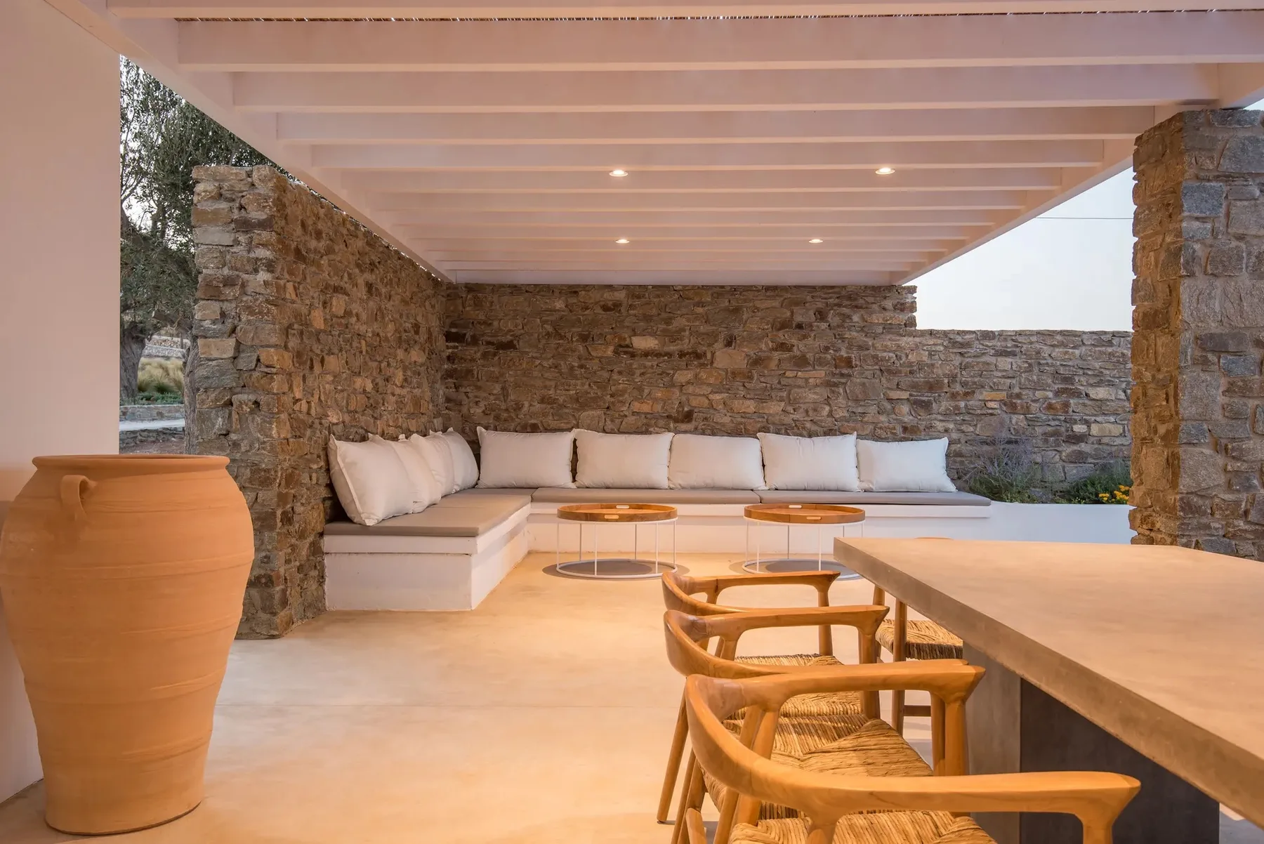 Covered outdoor lounge with built-in seating, a dining table, and stone walls.