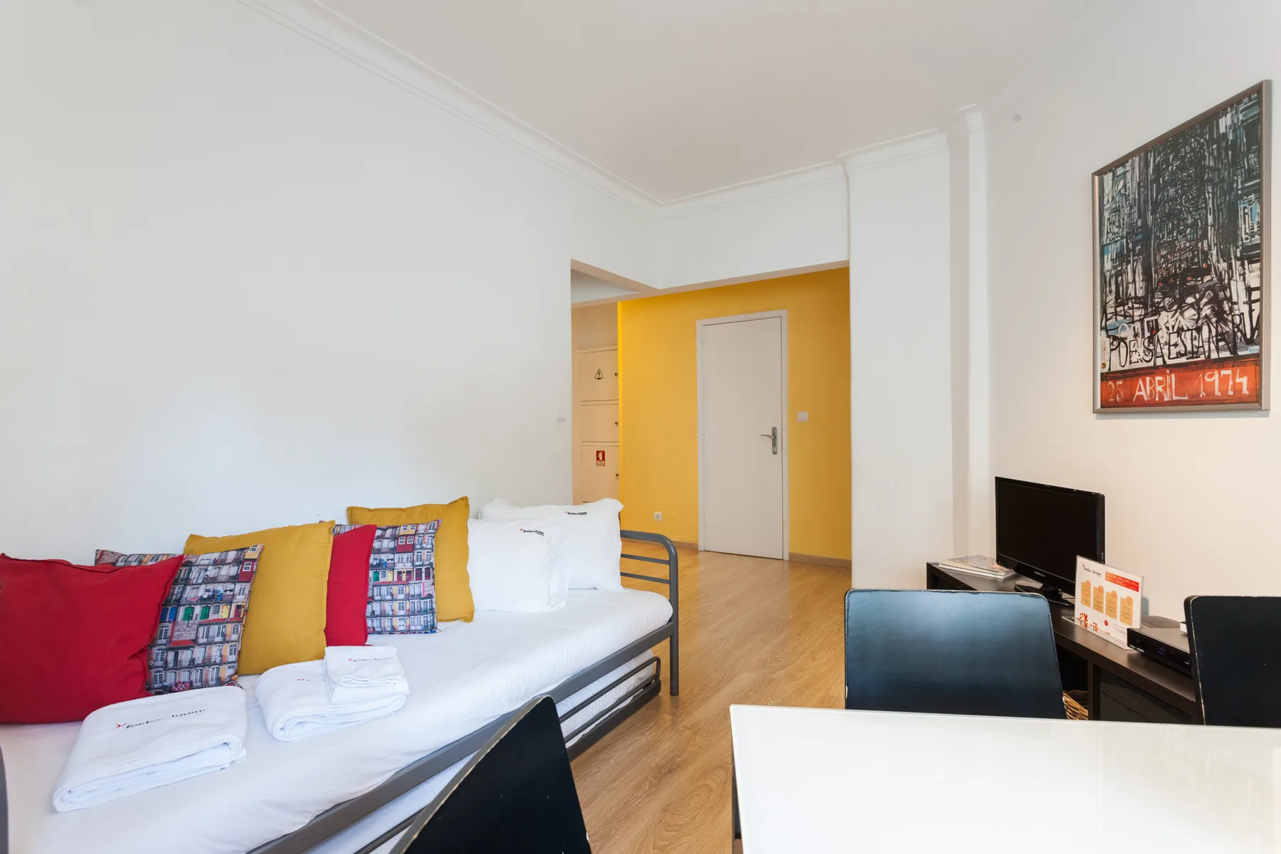 A bright room has a daybed with colorful cushions beside a desk and television.