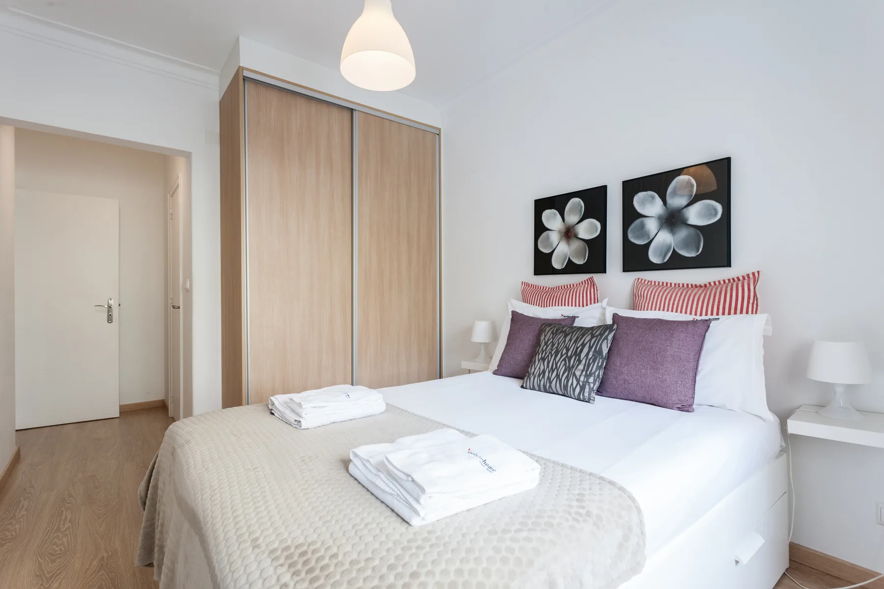 A bedroom with a neatly made bed, folded towels, and floral wall art.