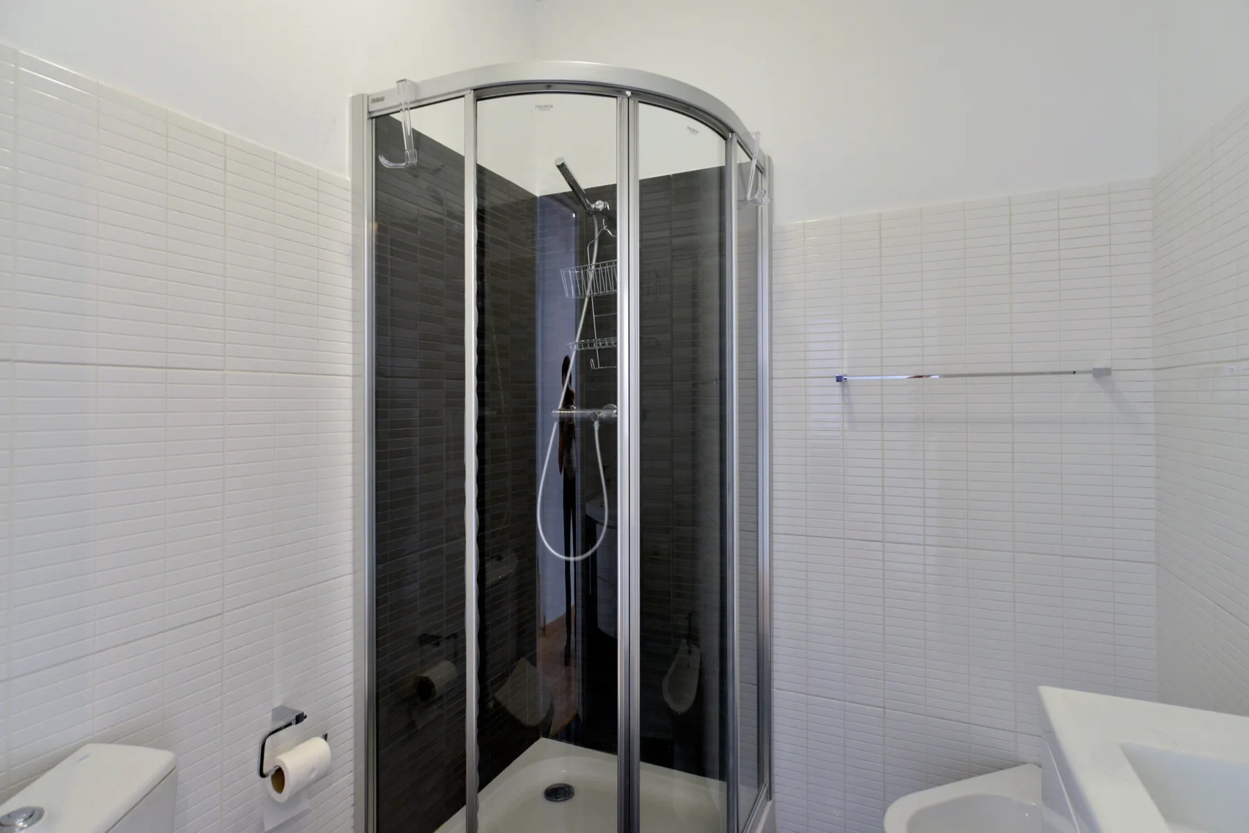 A bathroom with a curved glass shower and white tiled walls.
