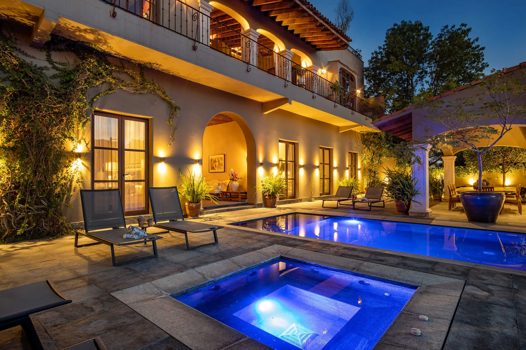An illuminated courtyard pool sits beside a two-story building and outdoor seating.