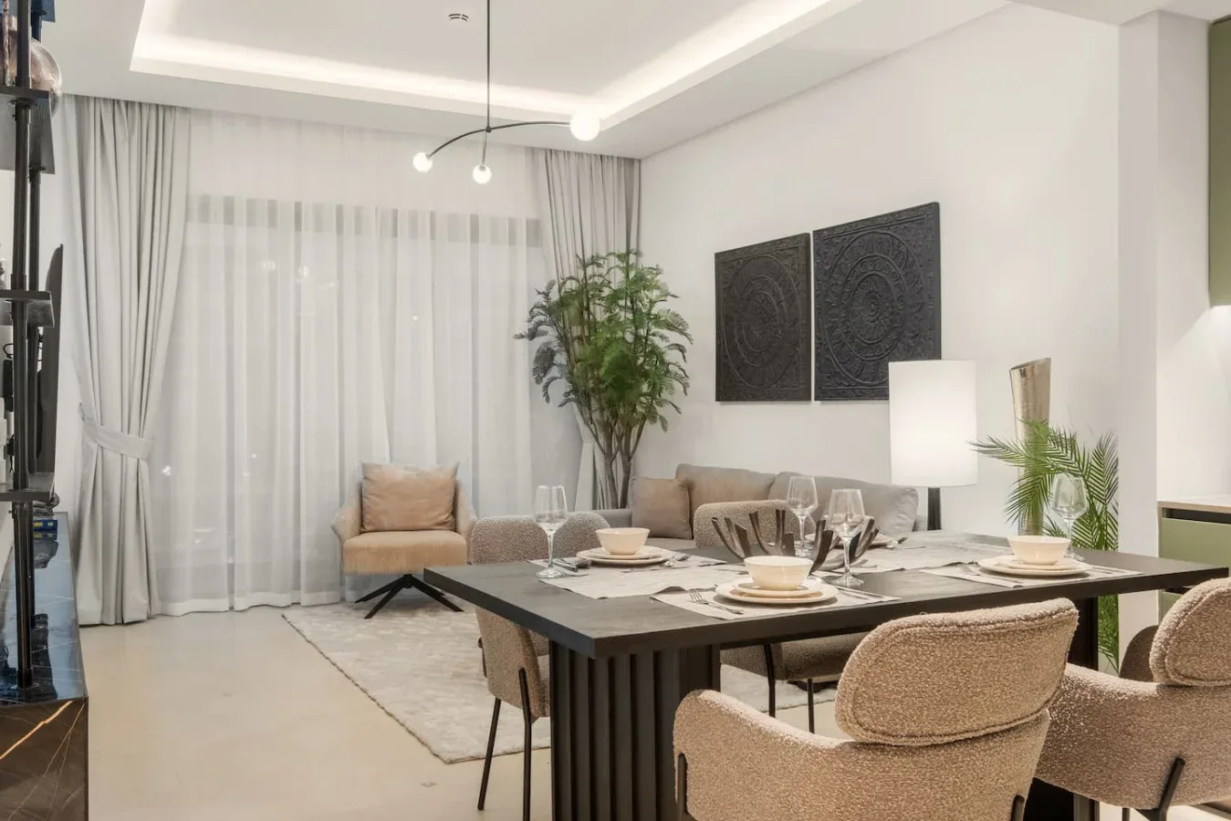 Un comedor con la mesa puesta, asientos tapizados y grandes ventanas con cortinas.