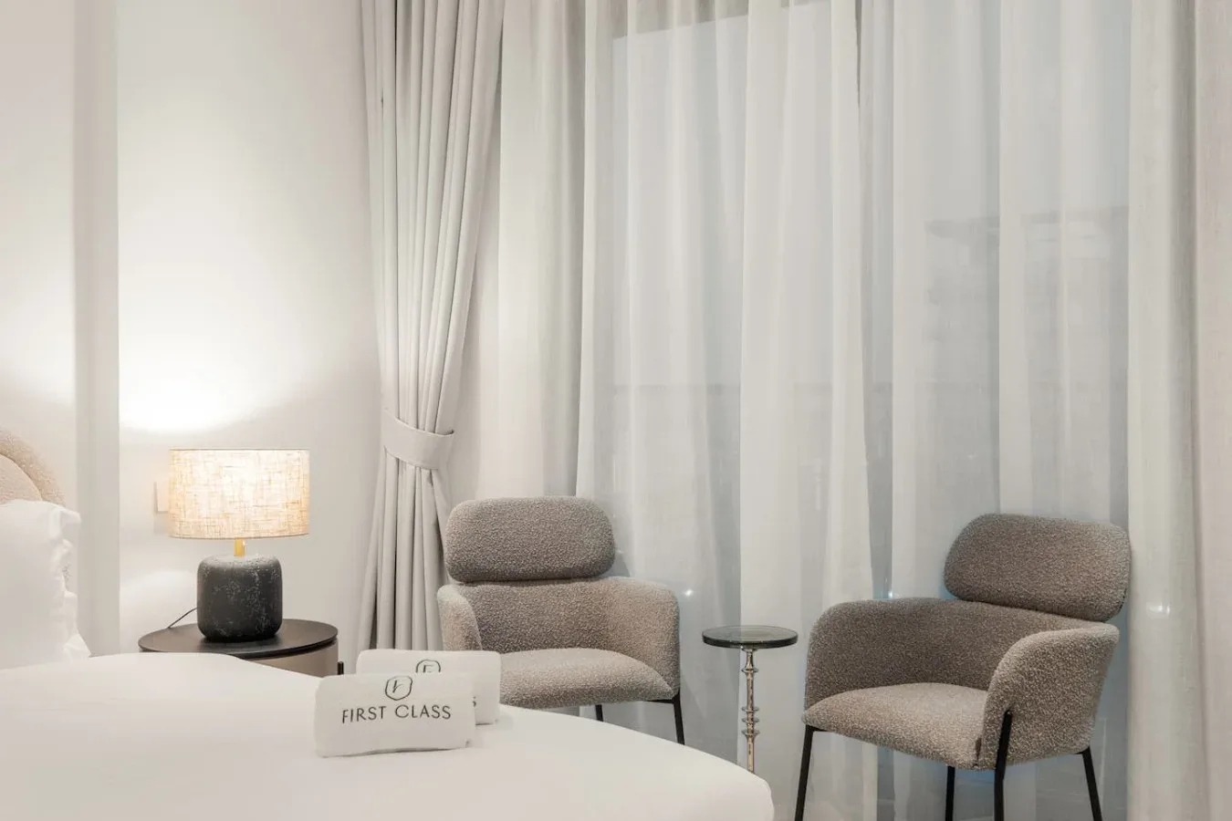 Un dormitorio con una cama, dos asientos tapizados y cortinas translúcidas.