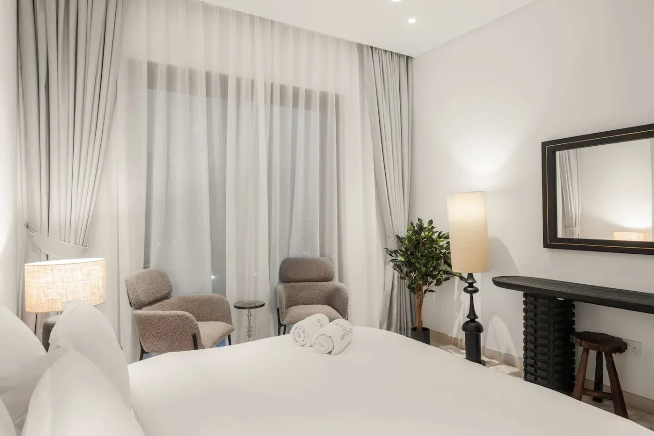 Un dormitorio con una cama, dos sillones y cortinas translúcidas frente a la ventana.