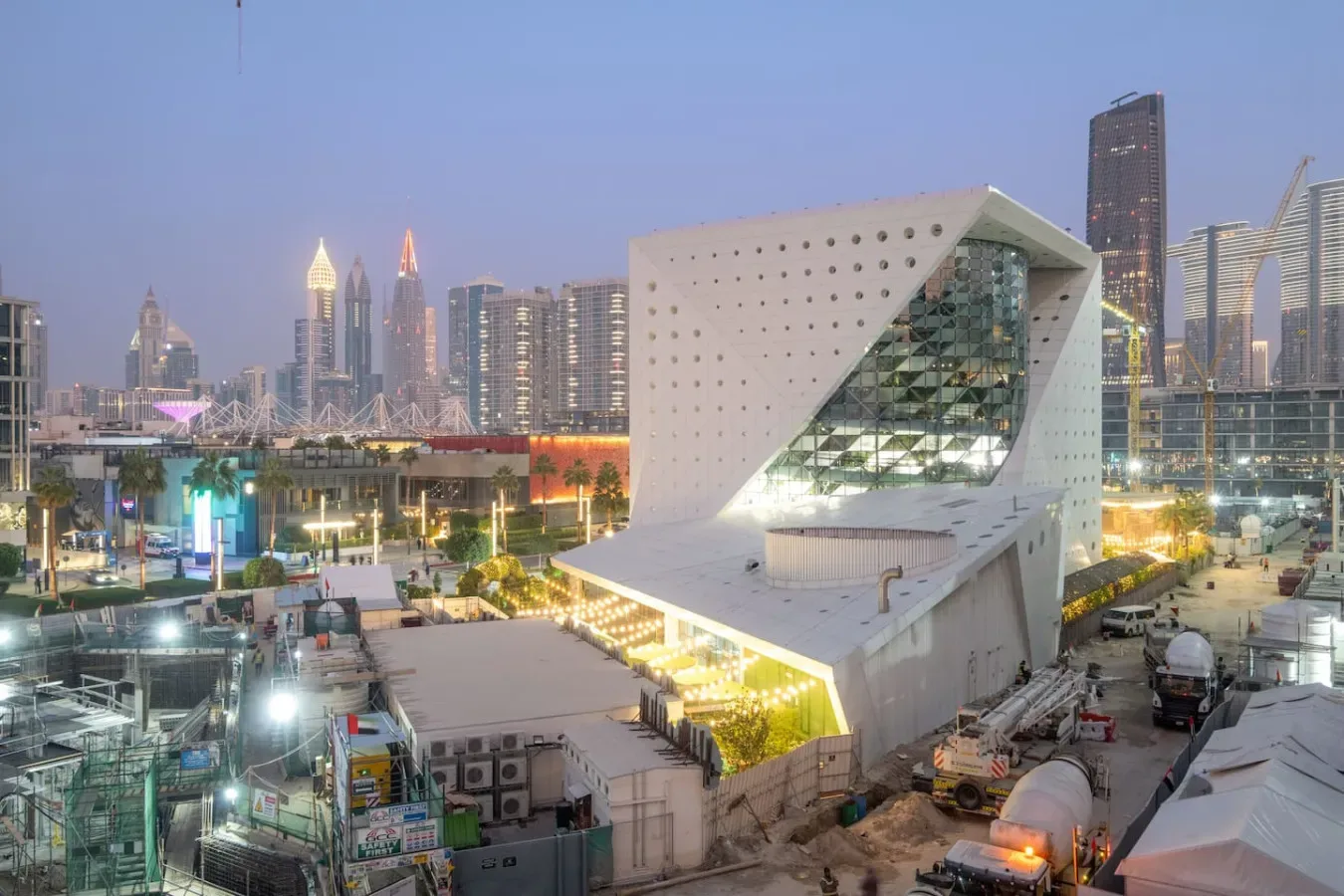 Un edificio moderno y angular se alza entre equipos de construcción y un horizonte urbano al anochecer.