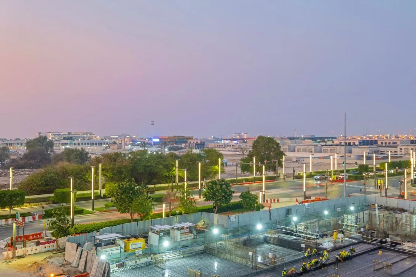El perfil urbano domina una vía ajardinada y una obra de construcción iluminada.