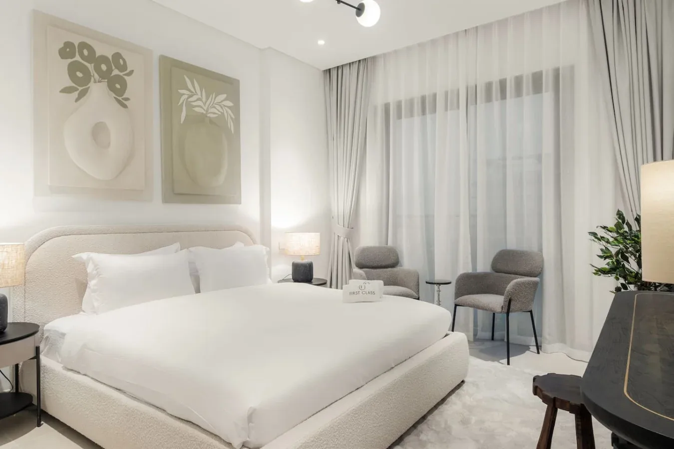 Un dormitorio luminoso con una cama blanca, arte botánico enmarcado y ventanas con cortinas largas.
