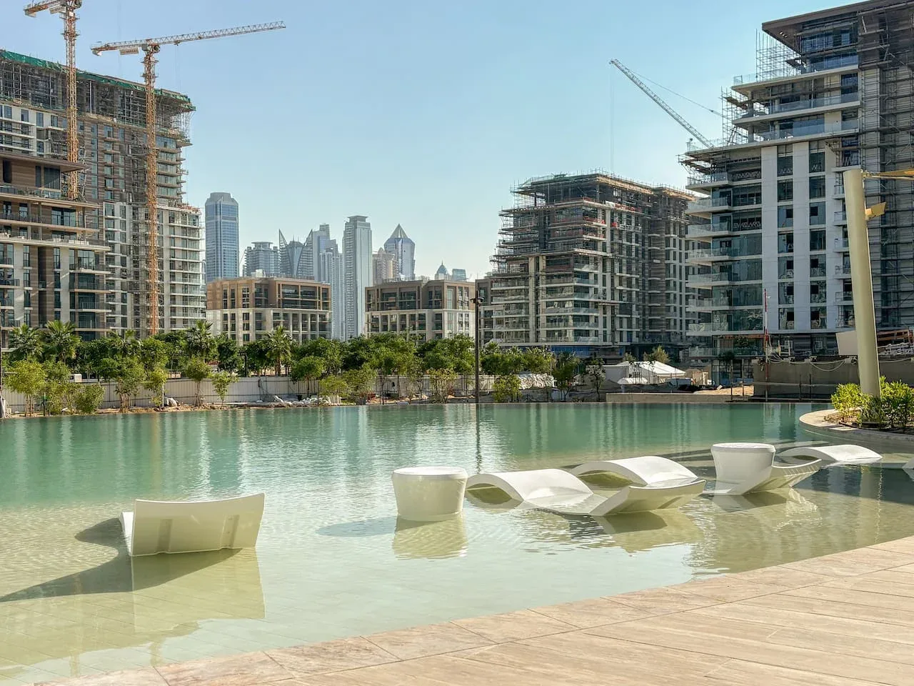 Una piscina poco profunda al aire libre está junto a una terraza de madera, con edificios al fondo.