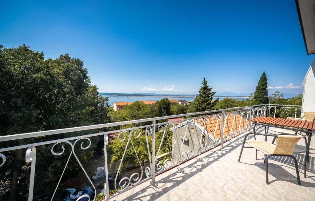 A sunlit balcony with outdoor seating overlooks trees and a distant stretch of water.