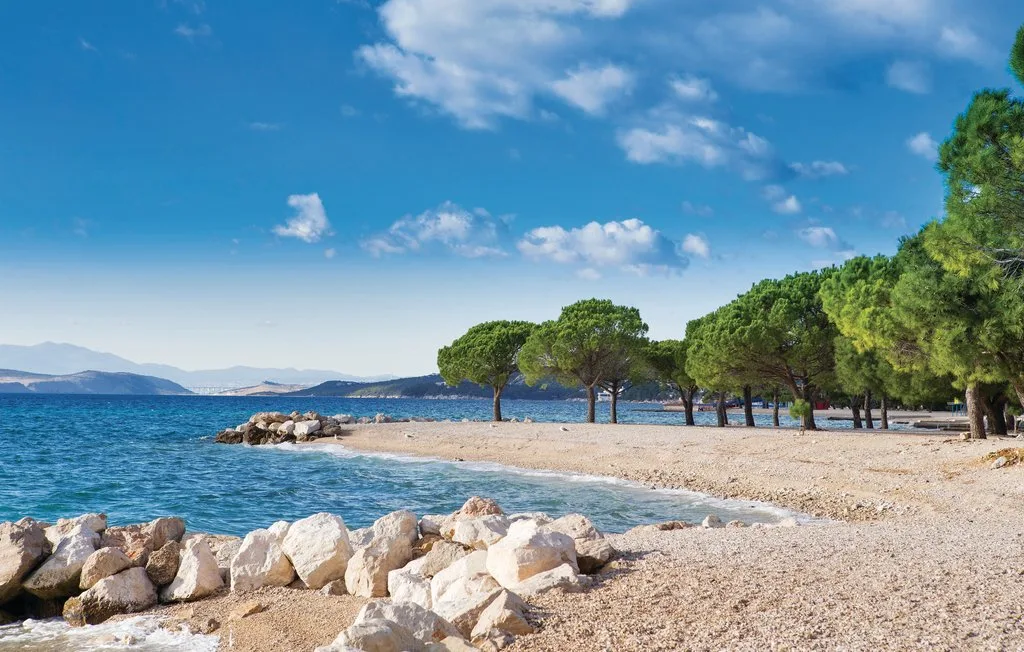 A pebble beach meets clear blue water beneath a line of trees.