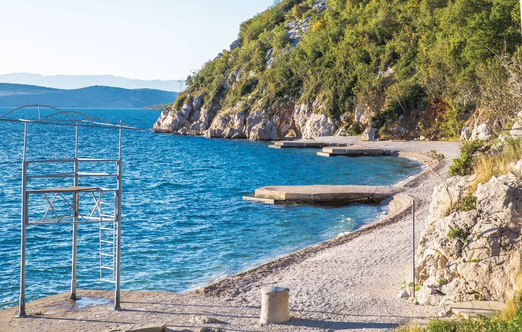 A small pebble beach beside clear blue water and a rocky, tree-covered shore.