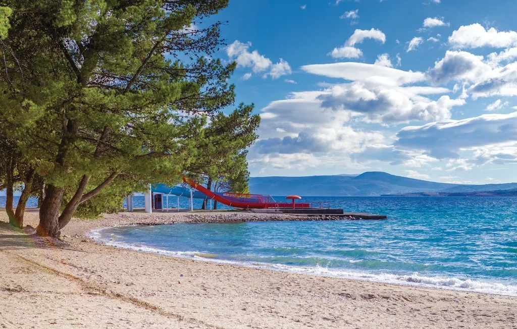 A sandy beach lined with pine trees faces blue water and distant hills.
