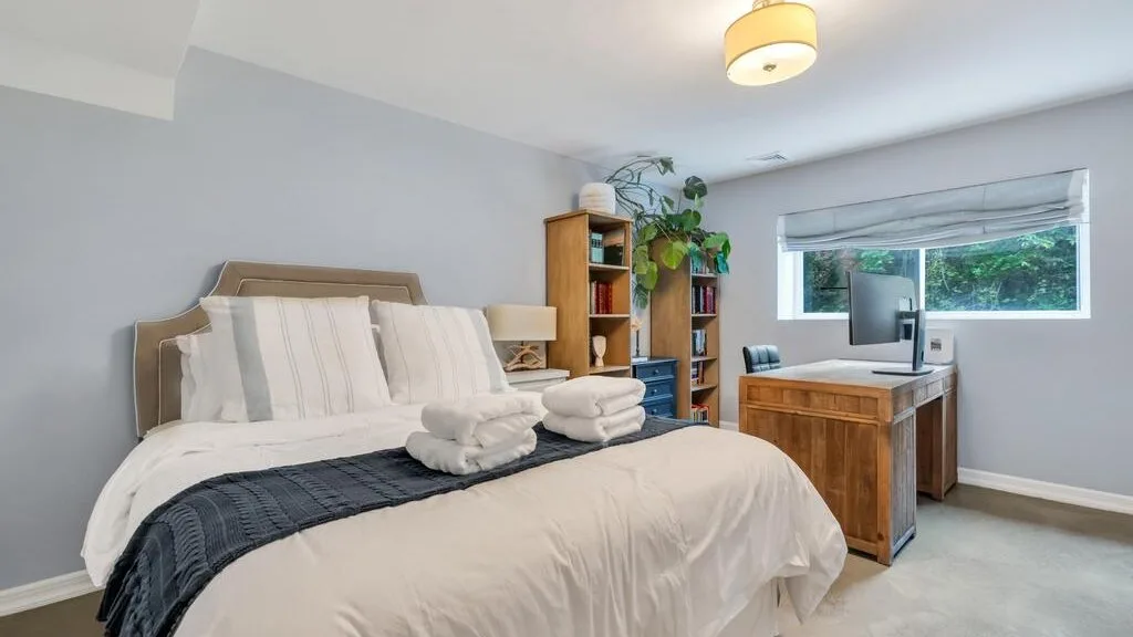 A bedroom with a bed, folded towels, and a wooden desk beside the window.