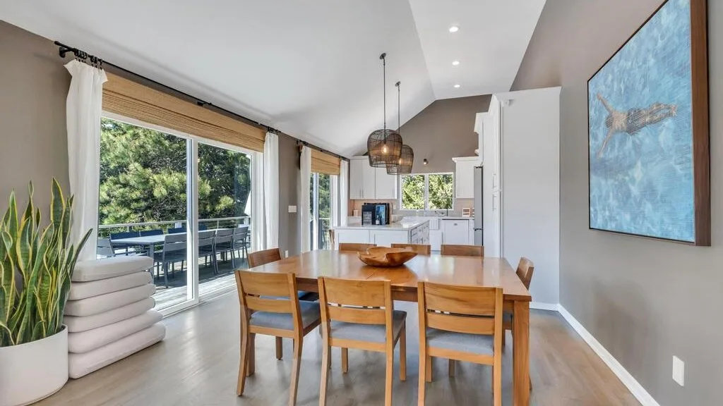 A dining area with a wooden table and chairs beside large windows.
