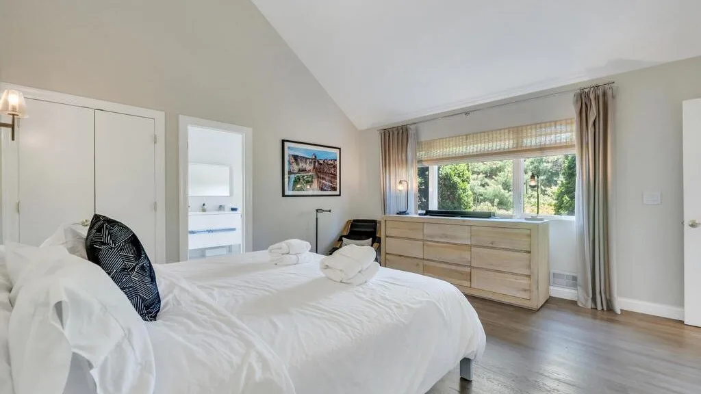 A bright bedroom with white bedding and a wide window overlooking greenery.