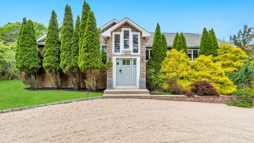 A house entrance faces a broad gravel driveway, with tall trees and shrubs along the front.