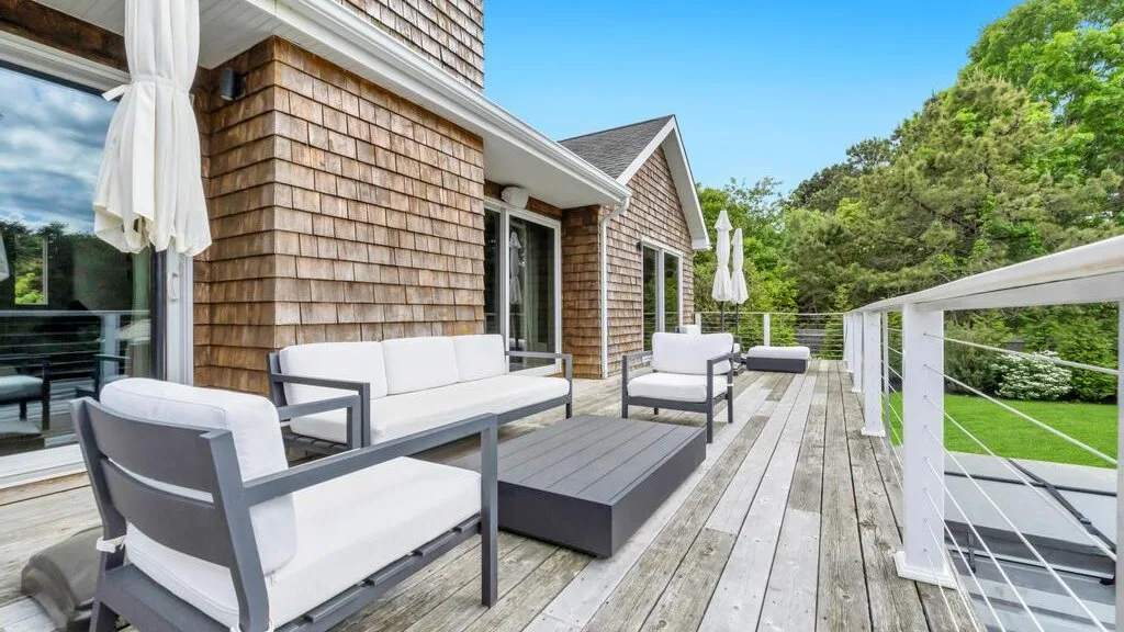 A wood-sided house opens onto a deck with cushioned seating and a green lawn.
