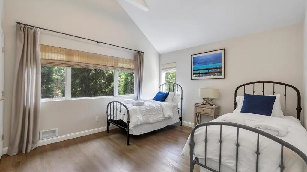 A bright bedroom with two beds, a large window, and framed wall art.