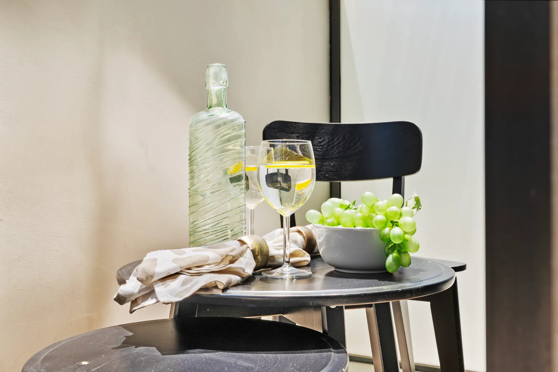 Una mesa pequeña sostiene una jarra de vidrio, una bebida, un paño doblado y un cuenco de uvas verdes.