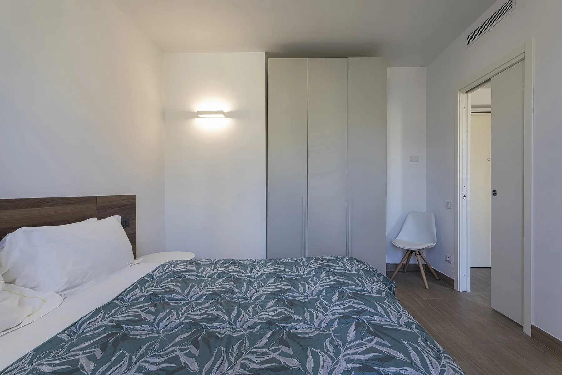 Un dormitorio con una colcha estampada, armarios empotrados y una silla pequeña.